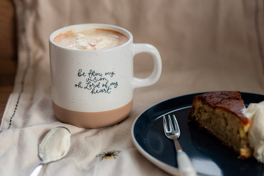 Minimalist Christian Mug with hand lettered words "be though my vision oh Lord of my heart" full ofhot chocolate with a plate with cake and some cream on a spoon.