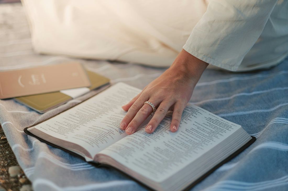 book lies open on a blanket with a woman's hand resting on it to imply reading and studying the Bible