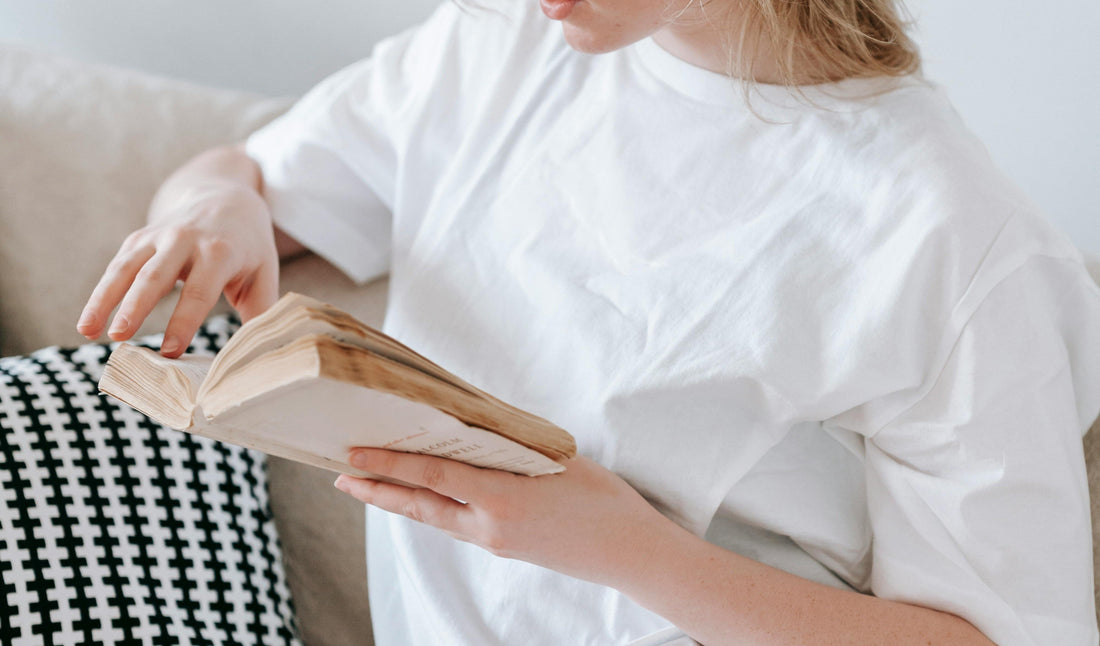 woman in a white t-shirt reading a paperback book on the sofa 