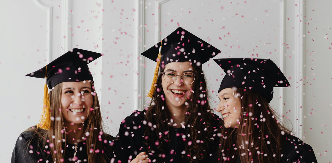 three white women in graduation cap and gowns surrounded by colourful confetti 