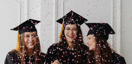three white women in graduation cap and gowns surrounded by colourful confetti 
