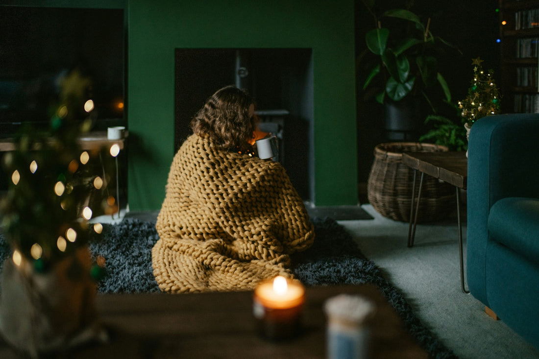 woman in a cosy room filled with fairy lights, candles, a hot cup of tea and wrapped in a blanket