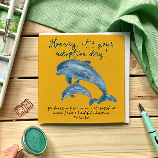 Christian adoption card with the words "Hooray it's your adoption day" as well as the scripture verse from psalm 16 "the lines have fallen for me in pleasant places" on a bright yellow background with dolphins
