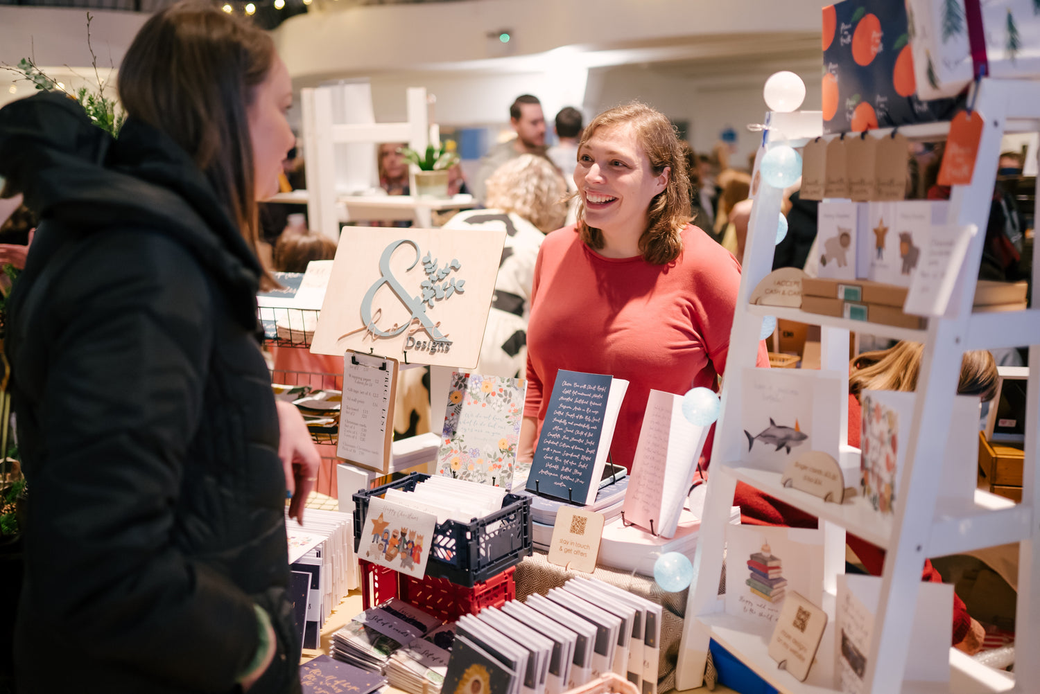 Photo of Anna Hamill at a craft fair chatting to a customer, with her stall of journals and cards in front of her.