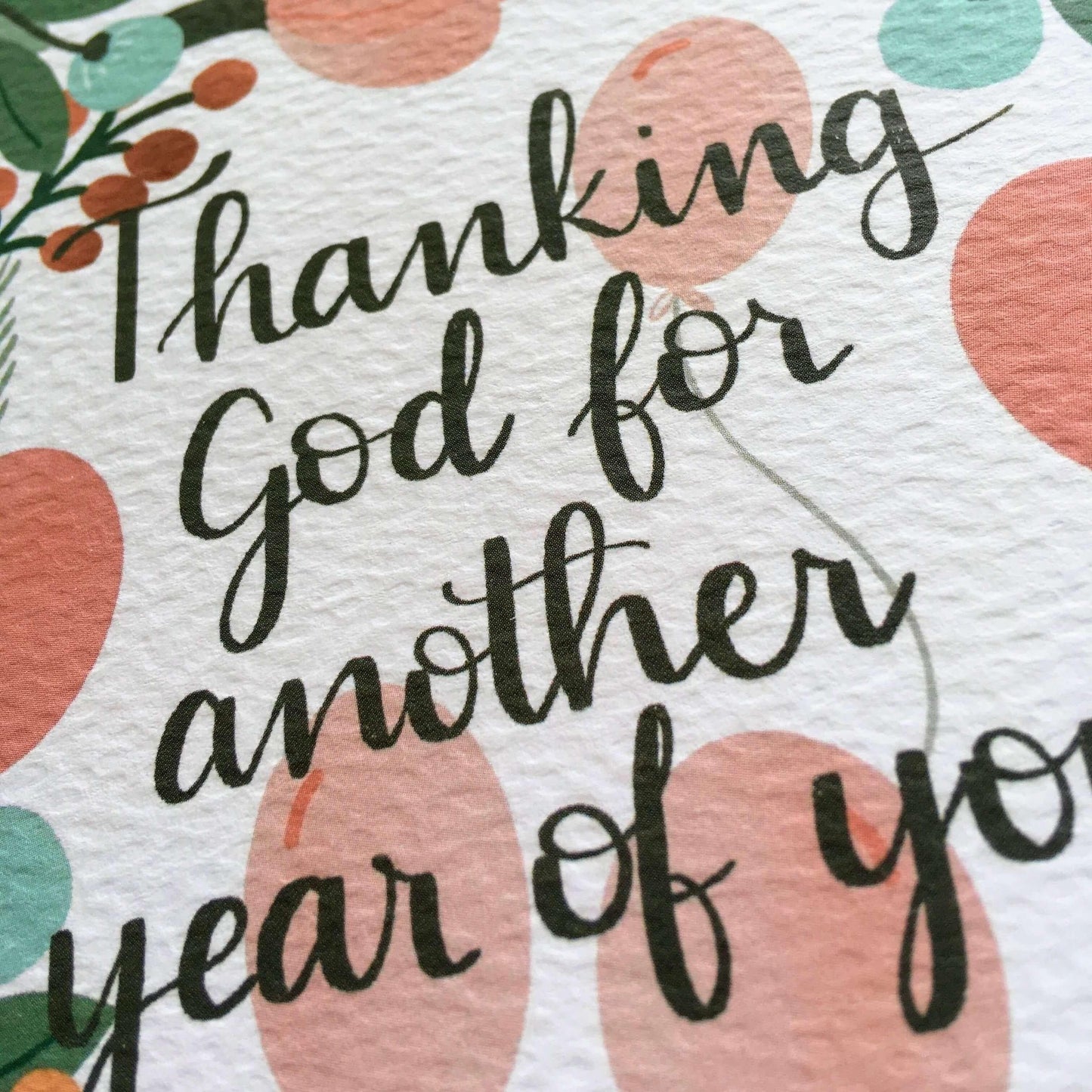 Close up on Christian birthday card with teal, mustard and pink flowers and balloons. Black hand lettered text saying thanking god for another year of you.